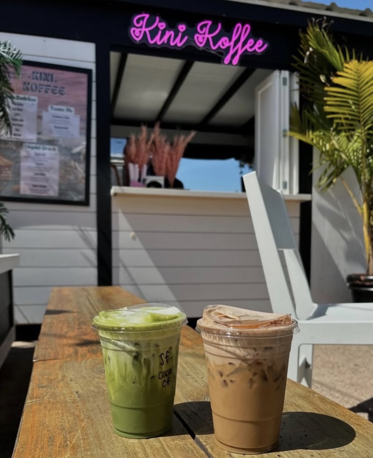 Two iced beverages, one green and one brown, on a wooden table outside a cafe with pink sign reading 'Kini Roffe'.
