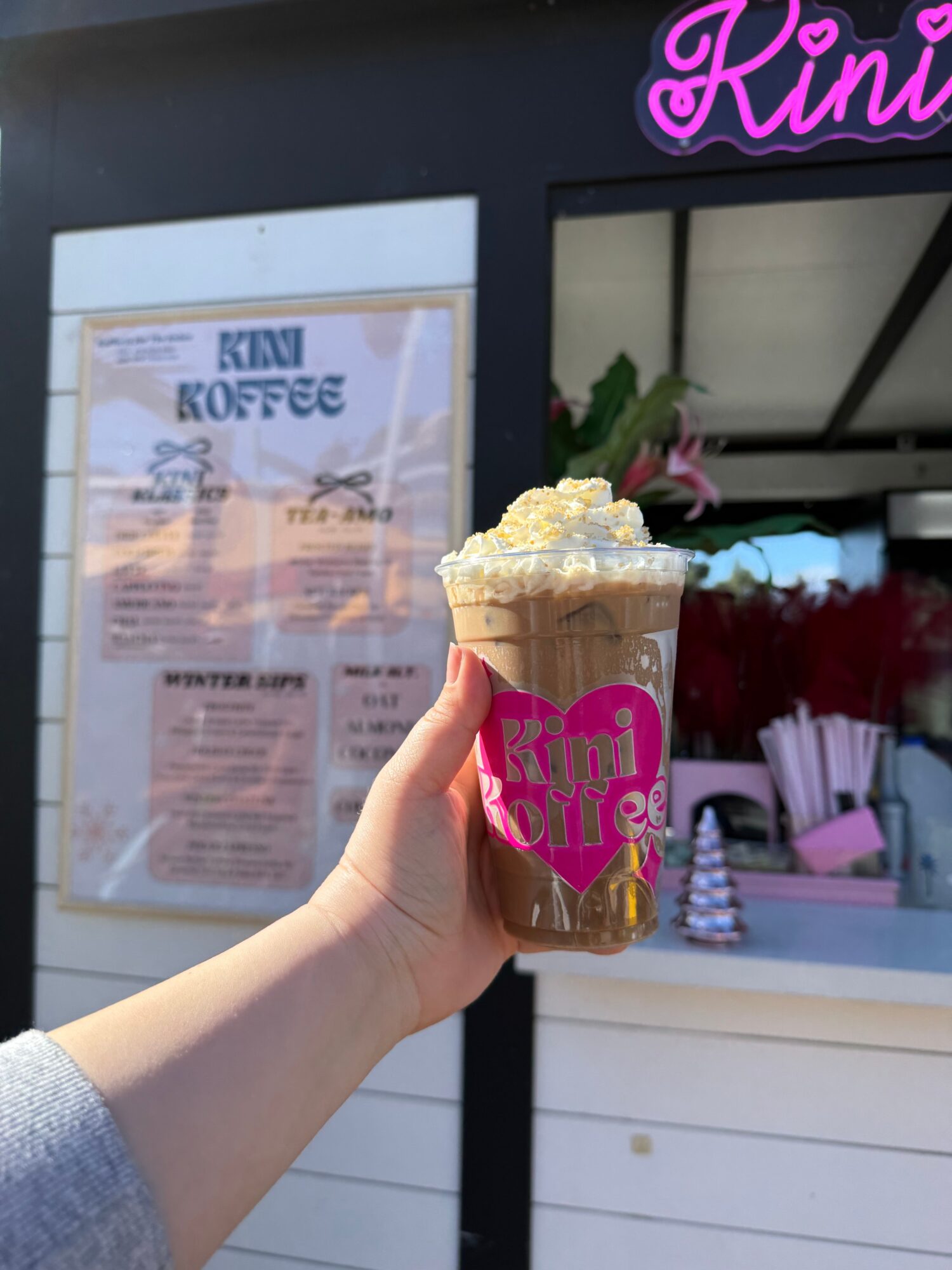 Person holding a coffee drink topped with whipped cream outside a cafe with pink signage and menu board.