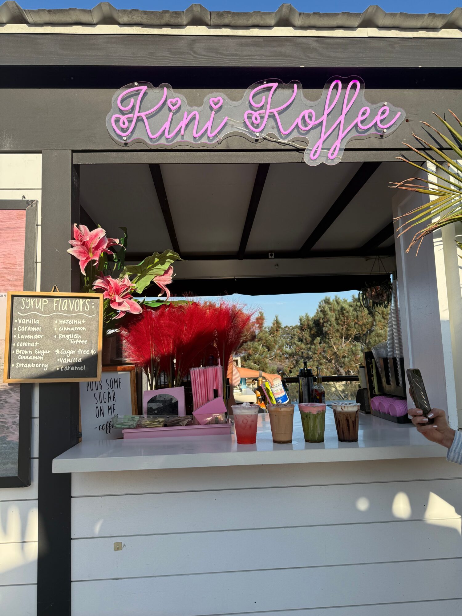 Kini Kaffee sign with pink neon lettering, outdoor counter with colorful drinks, and a menu board, sunny day.