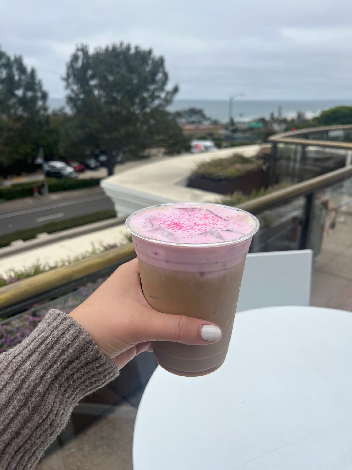 Person holding a pink-topped iced coffee near a window with trees and a road outside.
