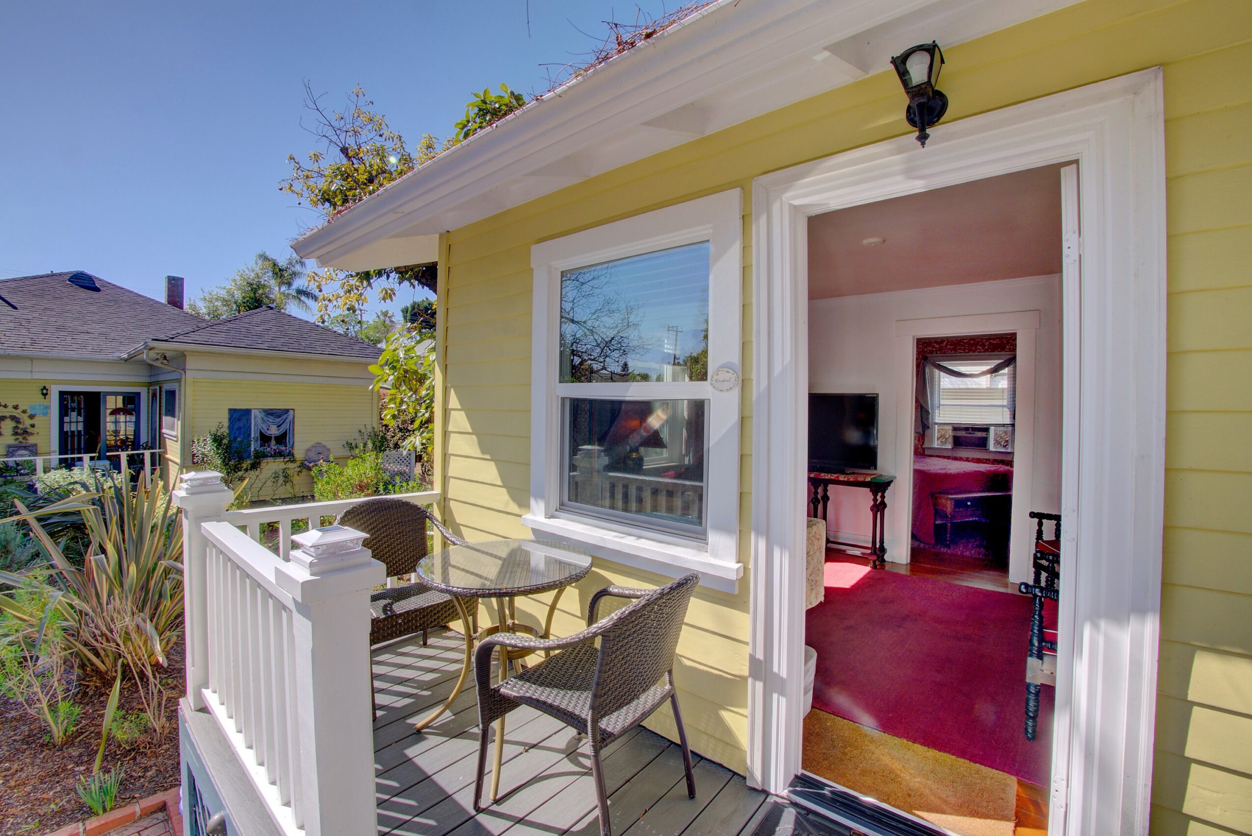 Yellow house with a porch, two chairs, and a table outside, view into a room with a TV and red carpet.