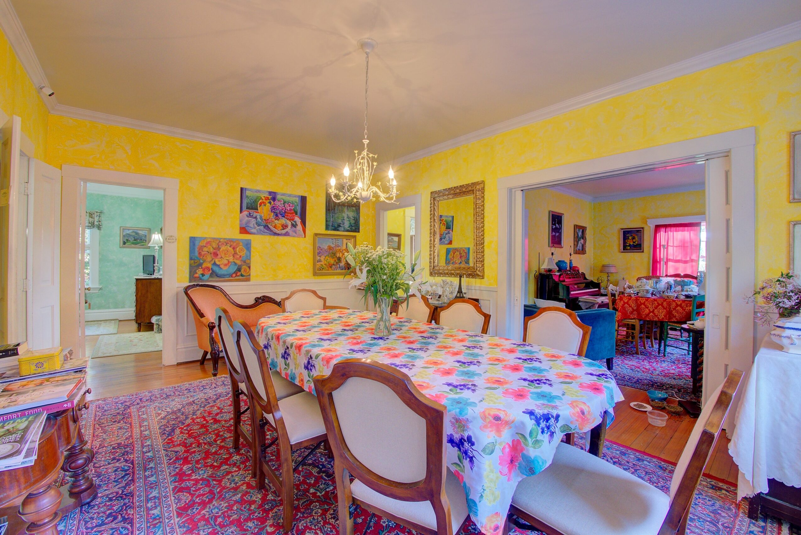 Dining room with a large table covered with a colorful tablecloth, surrounded by chairs, yellow walls, and artwork, chandelier overhead.