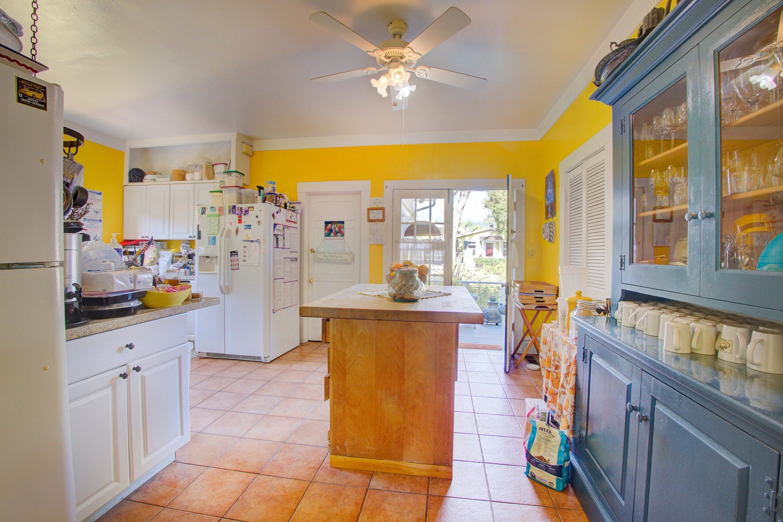Kitchen with yellow walls, white cabinets, blue hutch, and a small central island, sunlight through window, ceiling fan overhead.