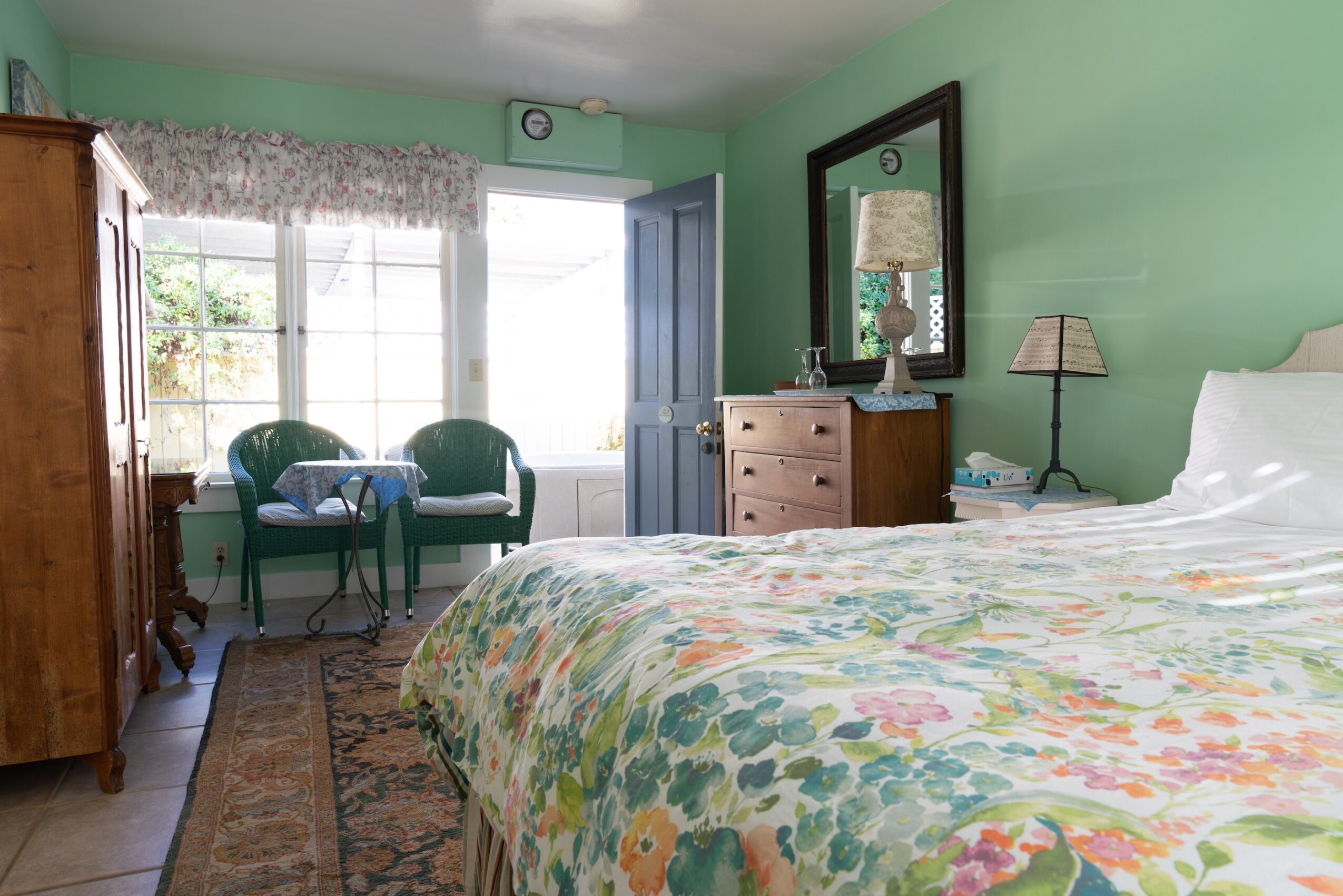 Bedroom with green walls, large window, bed with floral quilt, wooden dresser, mirror, and two chairs by window.