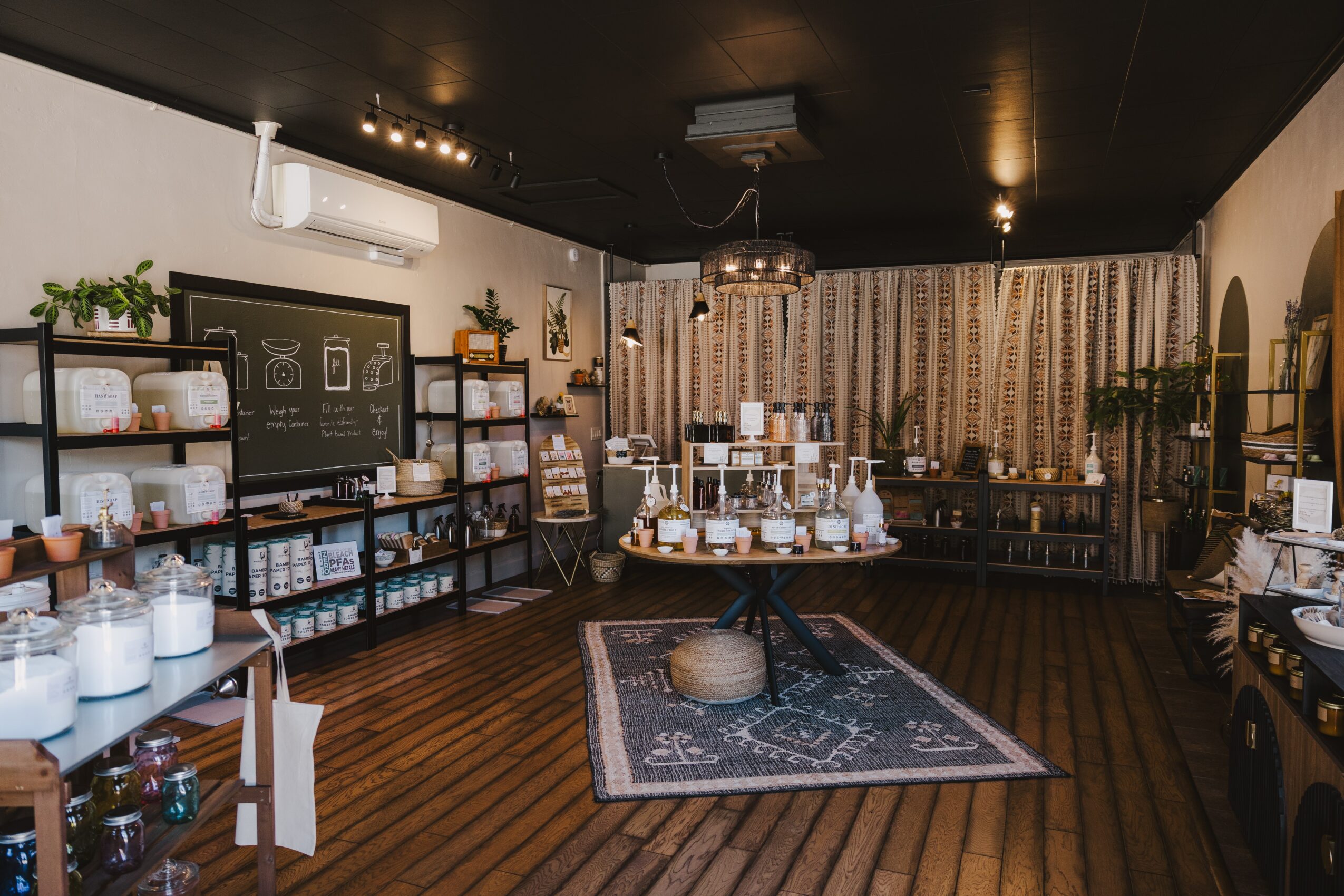 Interior of a store with shelves, a table with products, a rug, and decorative plants, ceiling lights, and curtains.