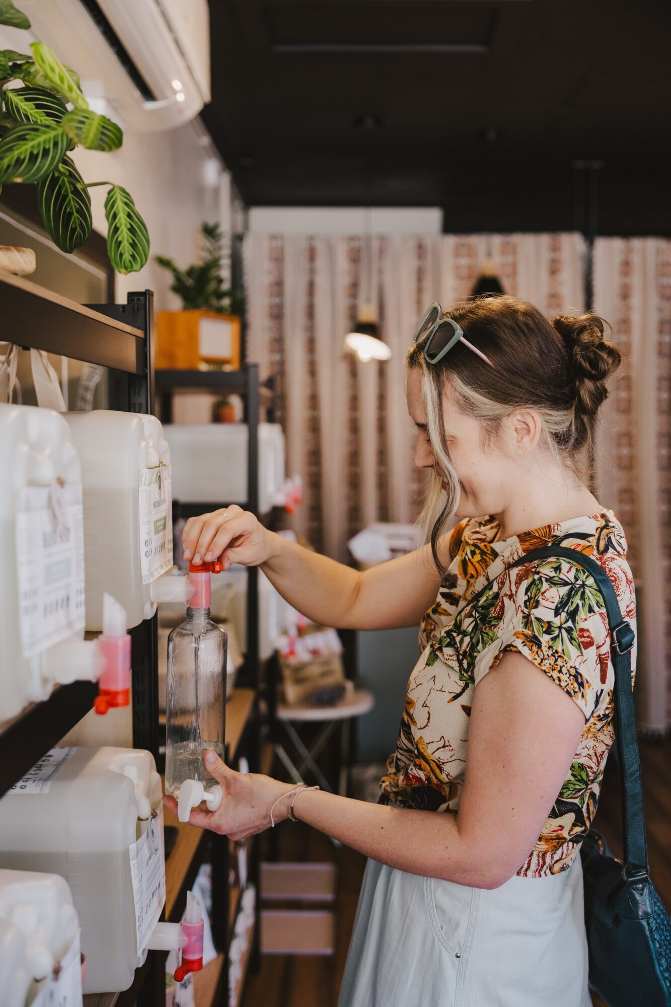 Young girl with glasses and a backpack shopping for items on a shelf in a store, wearing a floral top and white skirt.