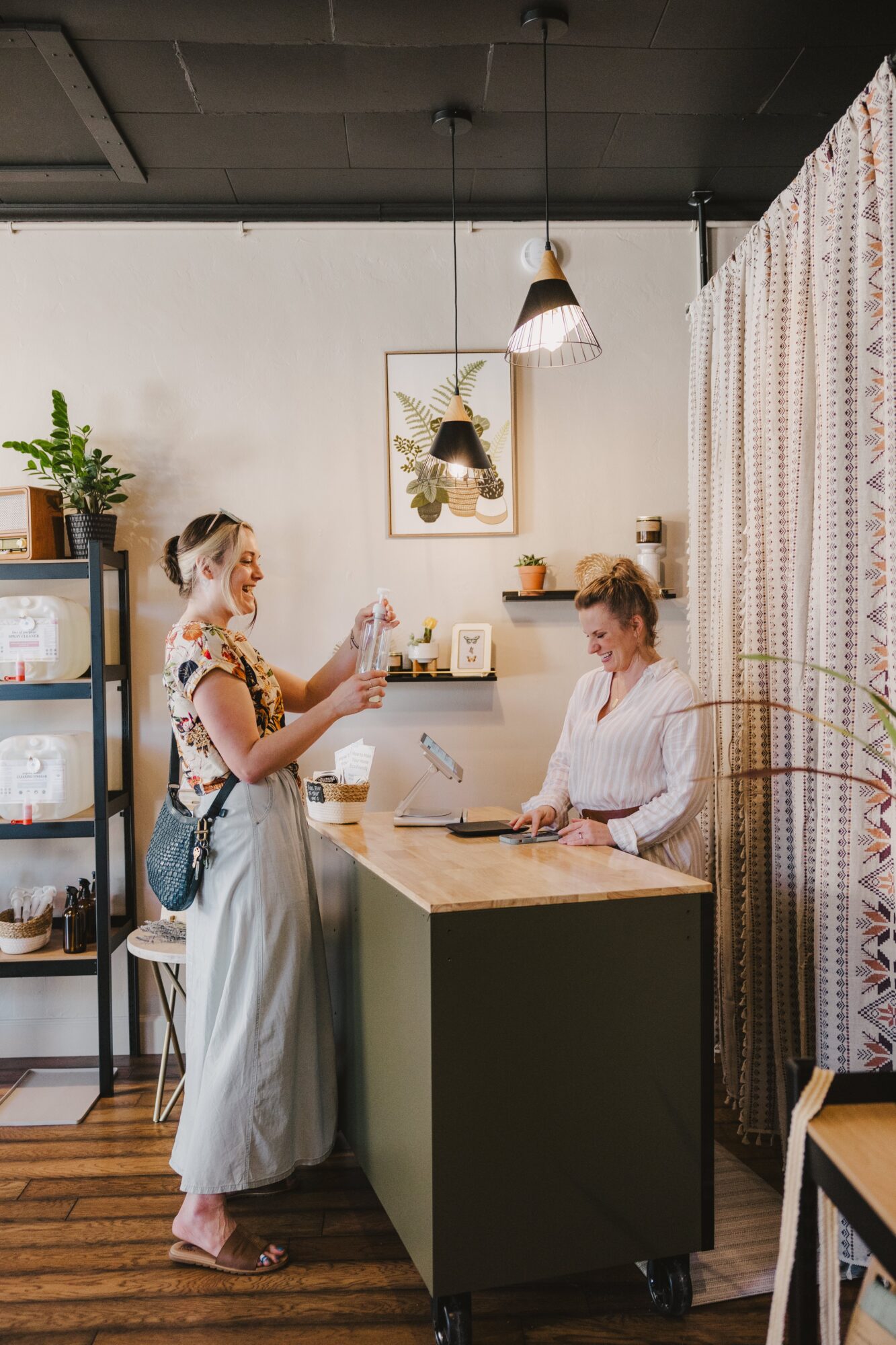 Two women in a cozy room, one standing and the other sitting at a counter, with plants and artwork in the background.