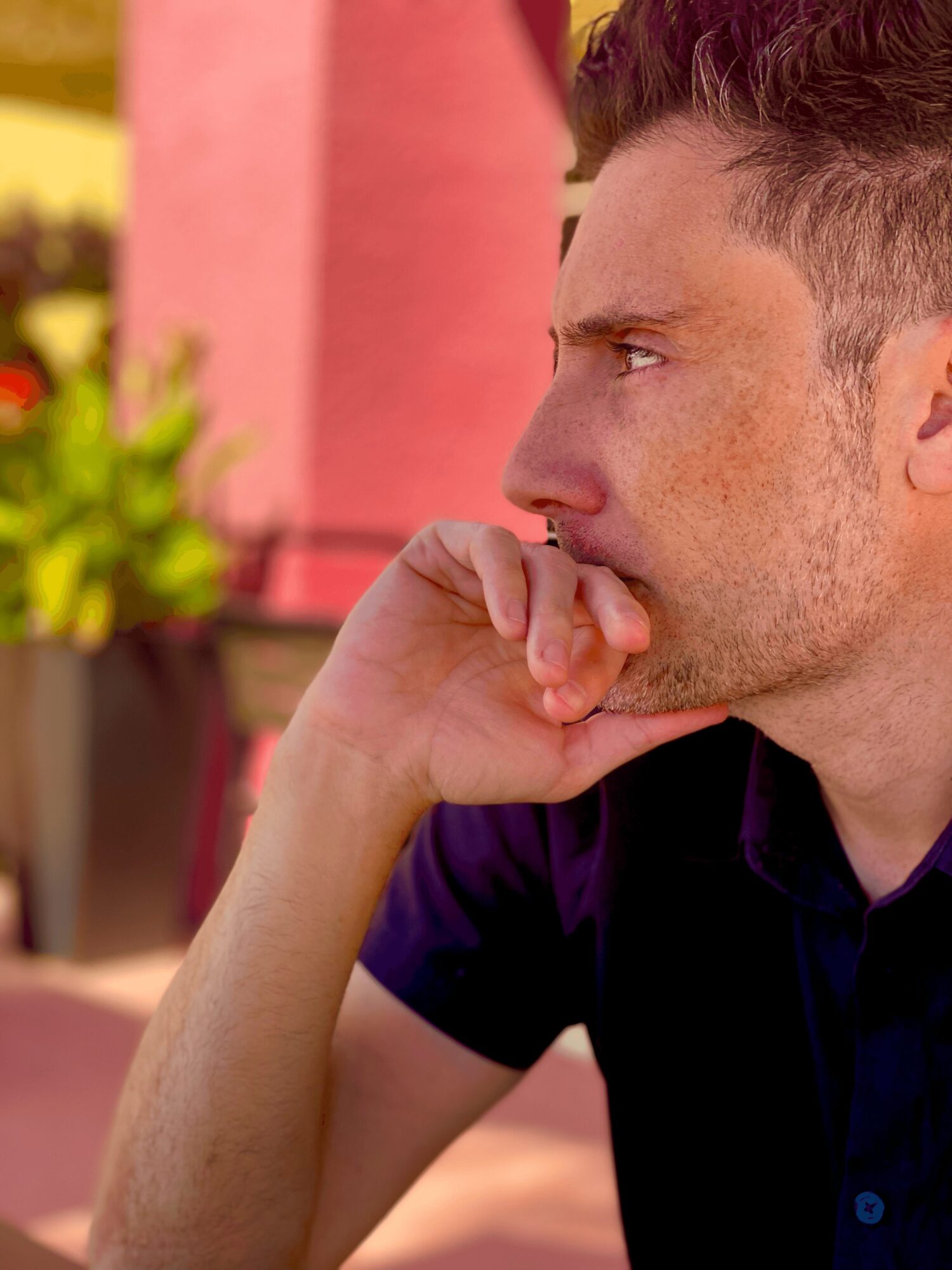 Close-up of a man with short hair, resting his chin on his hand, looking to the right outdoors.