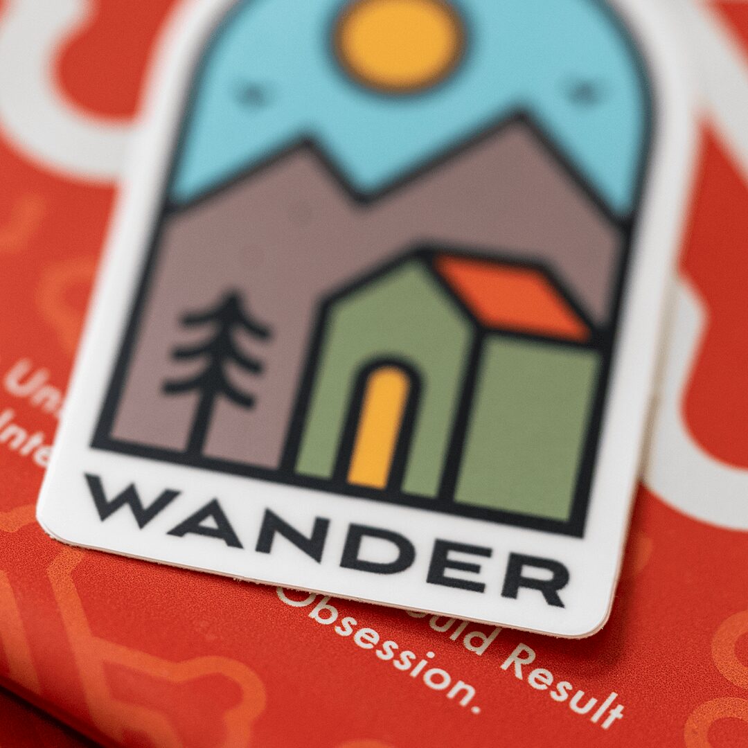 Sticker with a mountain, sun, tree, and house, labeled 'WANDER', on a red background.