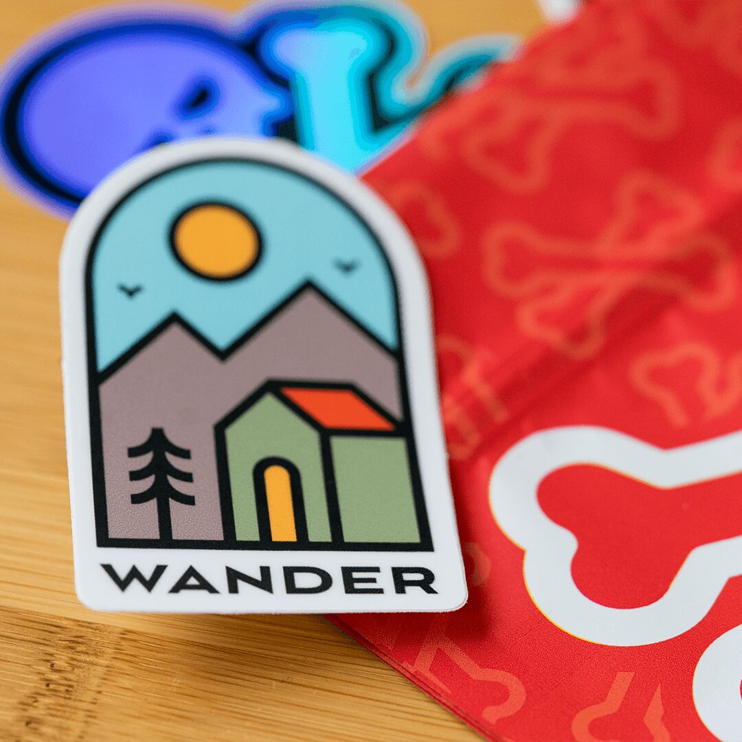 Sticker with a mountain, sun, house, and tree, labeled 'WANDER'.