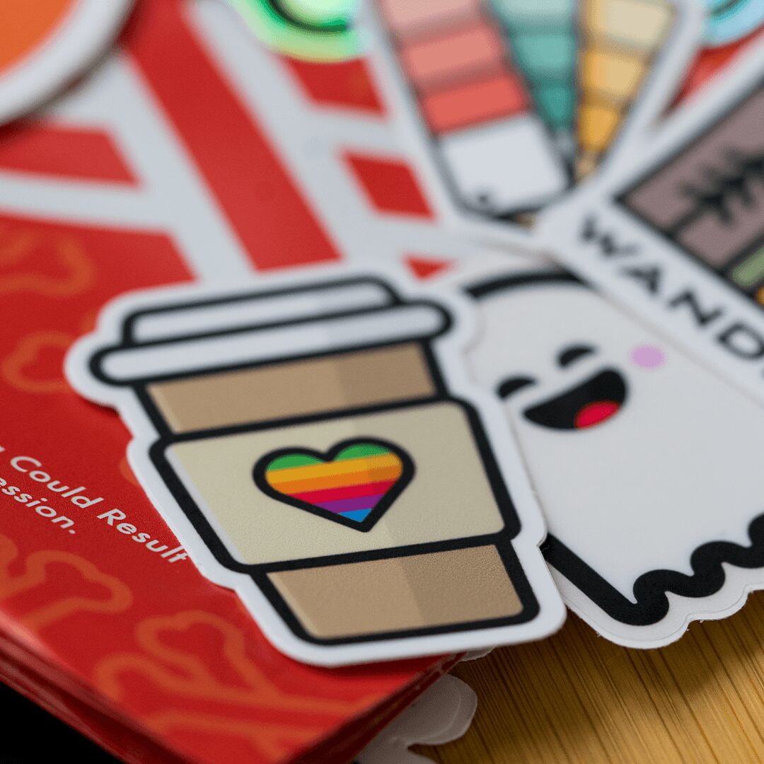 Sticker of a coffee cup with a rainbow heart, next to a smiling ghost, on a red background.