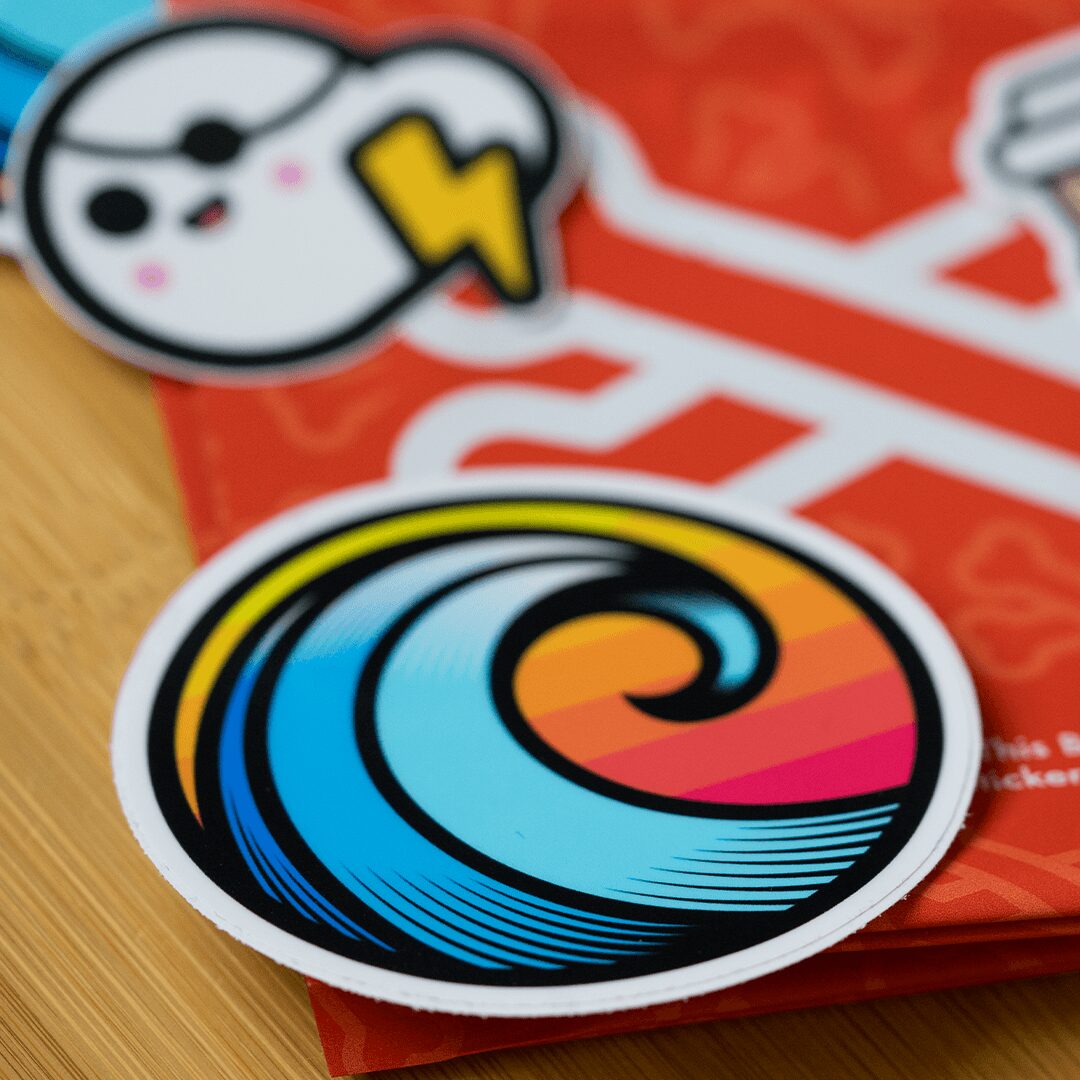 Two colorful stickers, one with a stylized face and lightning bolt, the other with a swirling wave design, on a wooden surface.