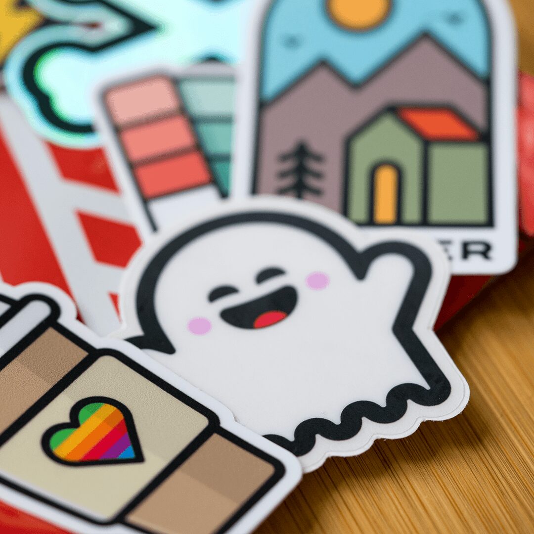 Collection of colorful stickers with various designs, including a smiling ghost, a rainbow heart, and outdoor scenery, on a wooden surface.