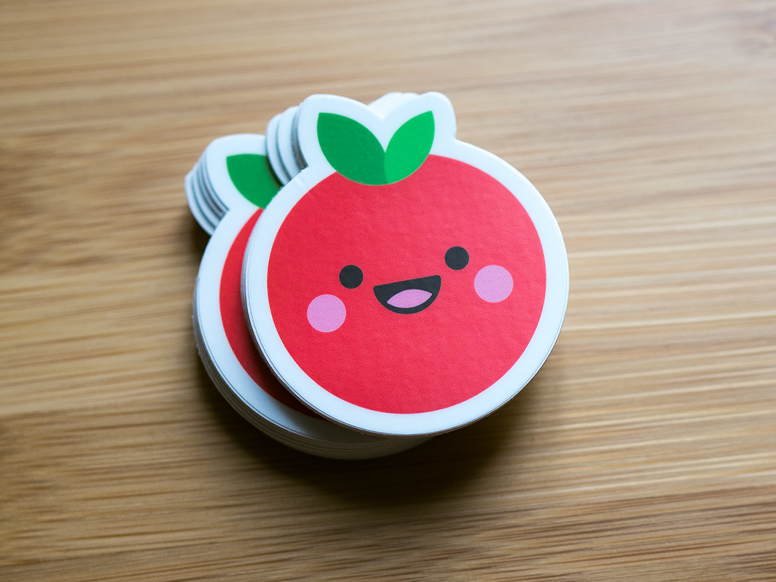 Stack of round stickers with a smiling, cartoon-style red fruit with green leaves on a wooden surface.