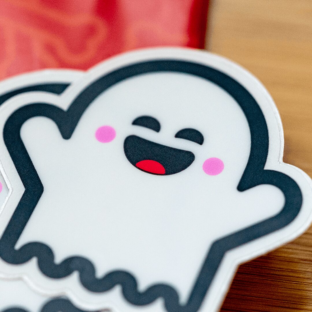 Cute cartoon ghost with a smiling face, pink cheeks, and waving hand, on a wooden surface.