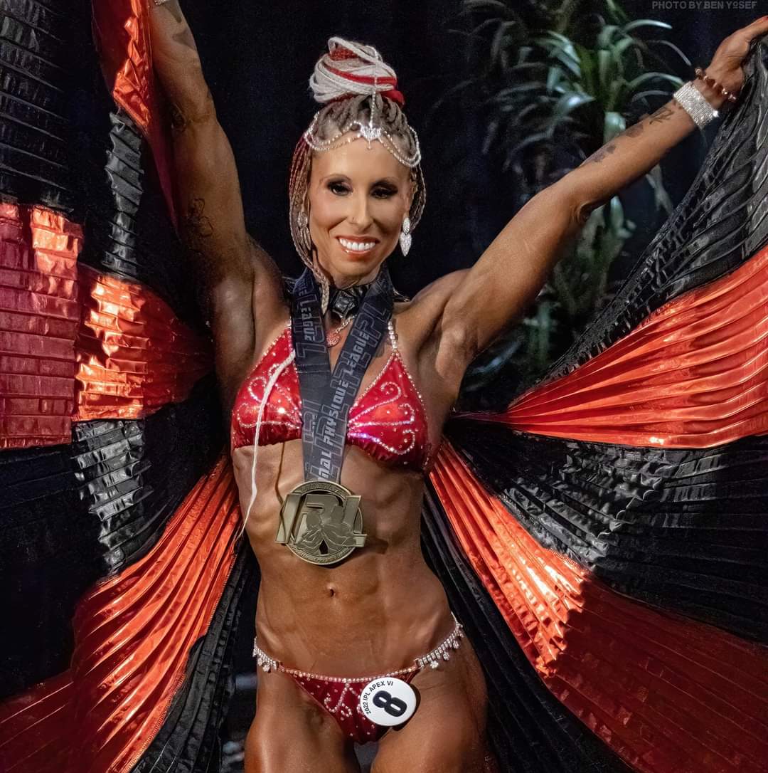 Woman in red and black costume with large wings, smiling, holding arms up, wearing a medal and sash, background with plants.