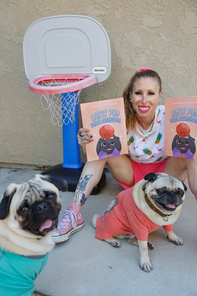 Woman smiling with two pugs, one in a teal shirt and the other in a pink shirt, outdoors near a small basketball hoop.