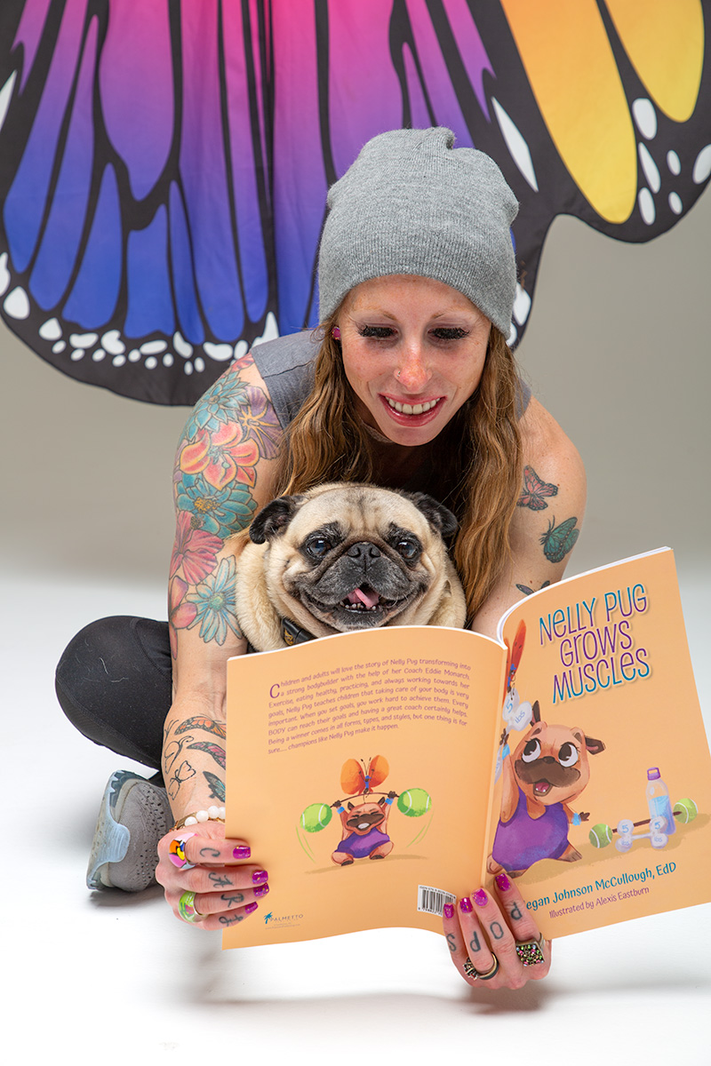 Woman with tattoos and gray beanie reading a children's book to a pug dog, colorful butterfly mural in background.