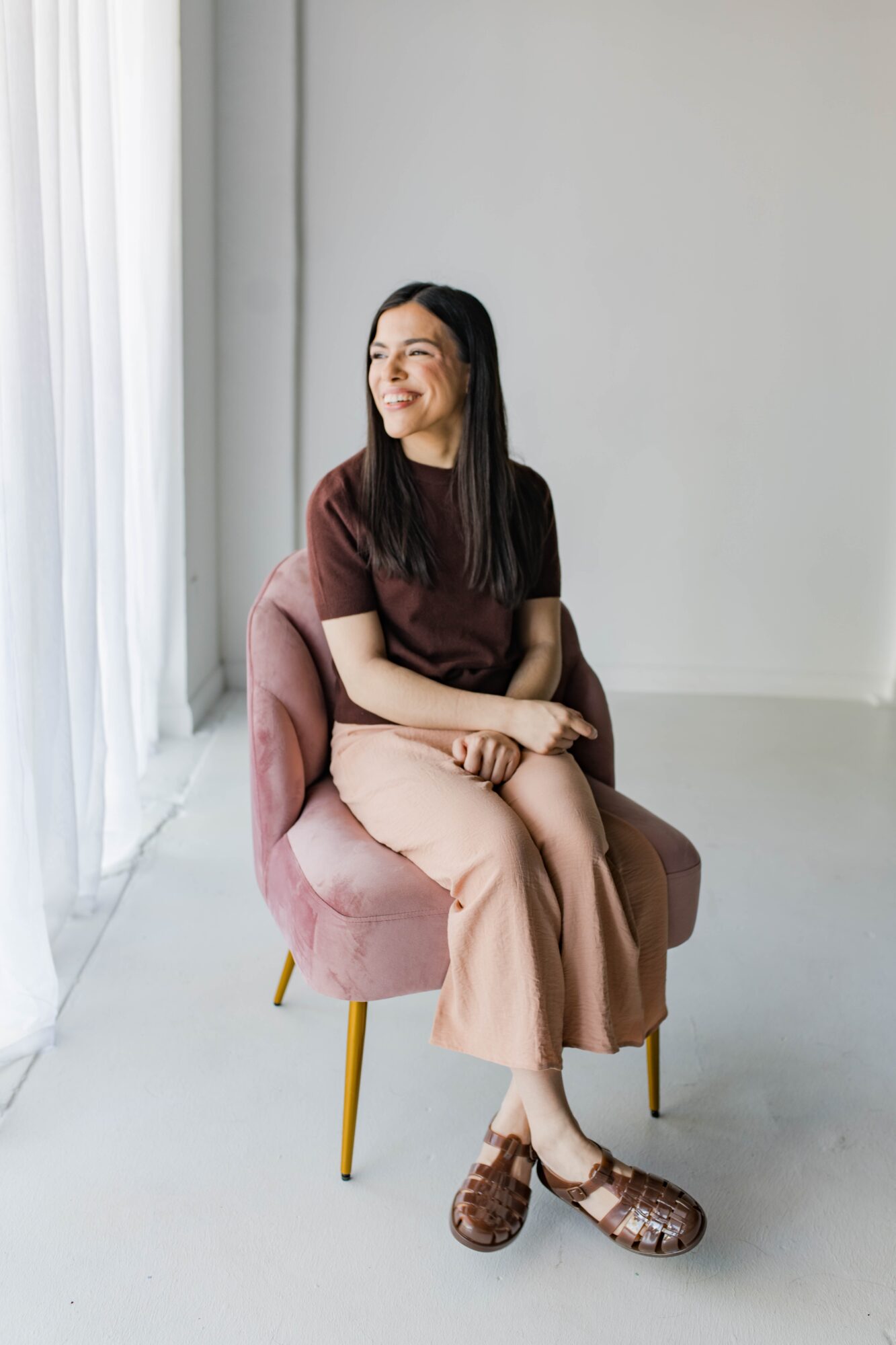 Woman sitting on a pink chair near a window, smiling and looking away, in a bright room.