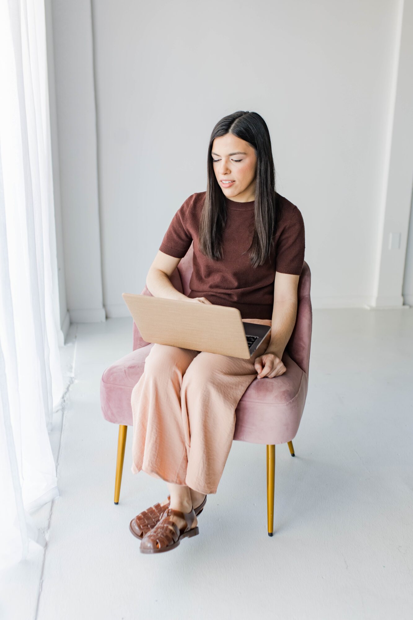 Woman sitting on pink chair, using laptop, in bright room with white walls and floor.