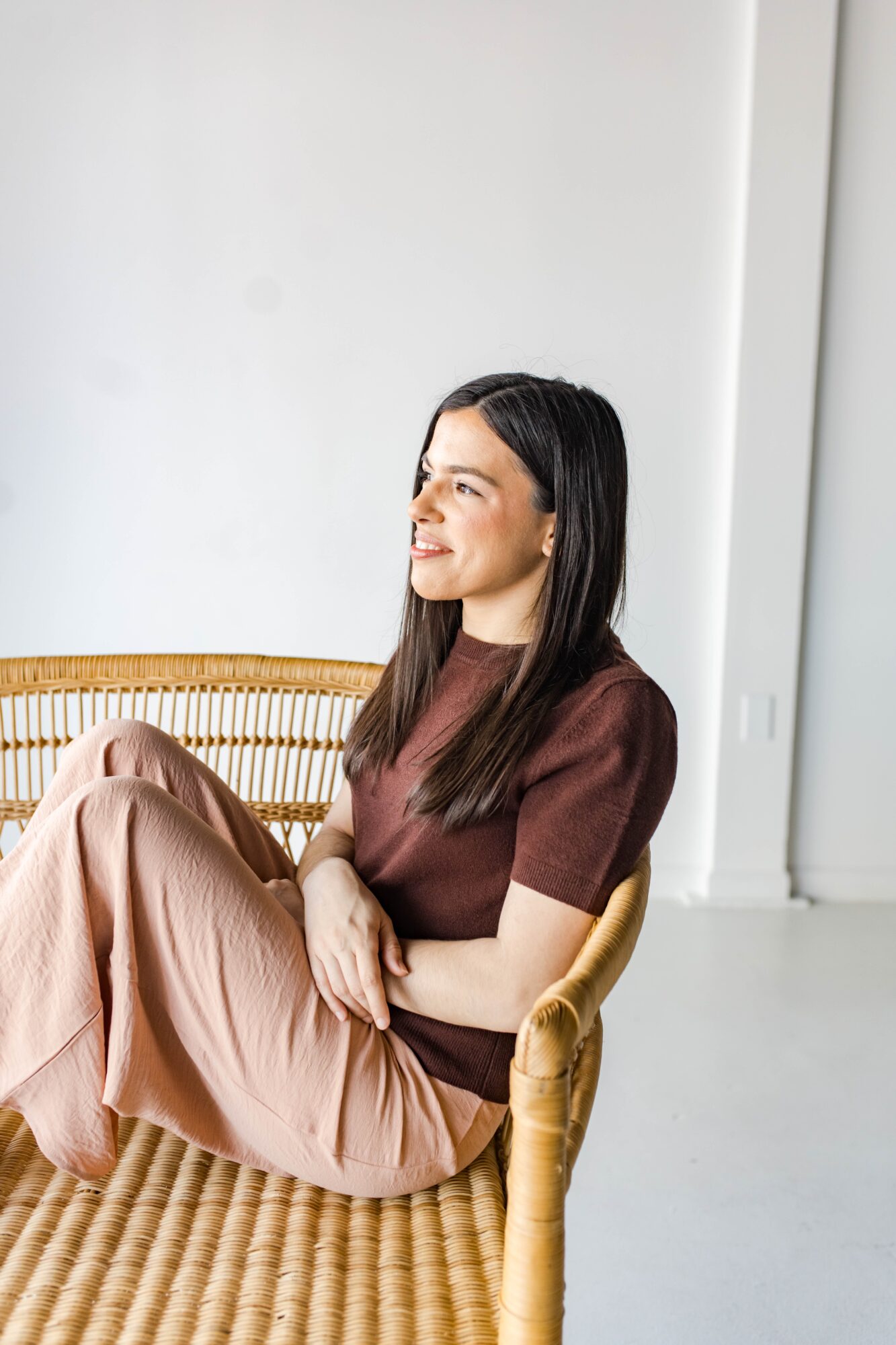 Woman sitting on a woven chair, smiling, with a plain white wall background.