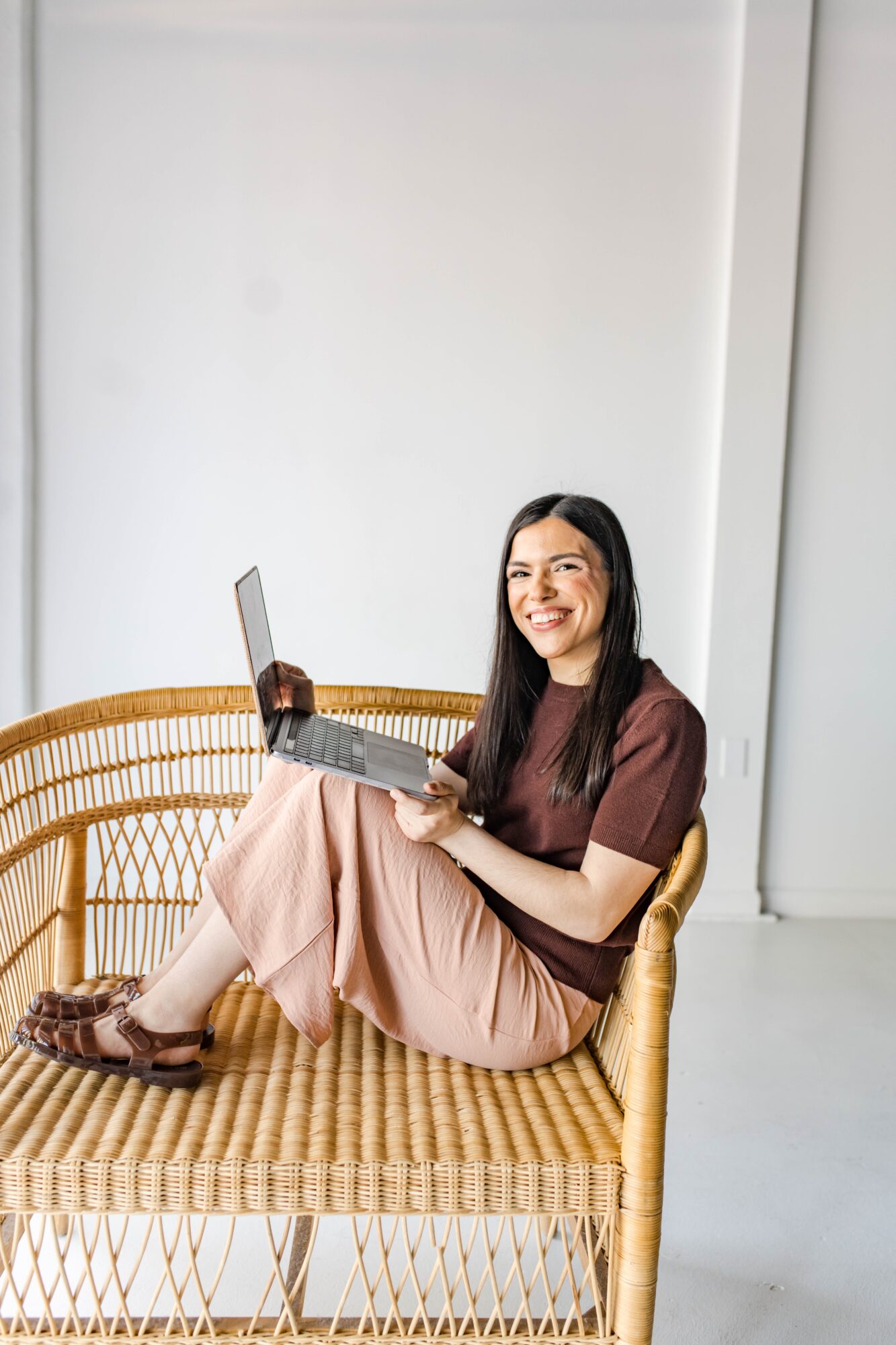 Woman sitting on a wicker bench, smiling, holding a laptop, in a bright room with white walls.