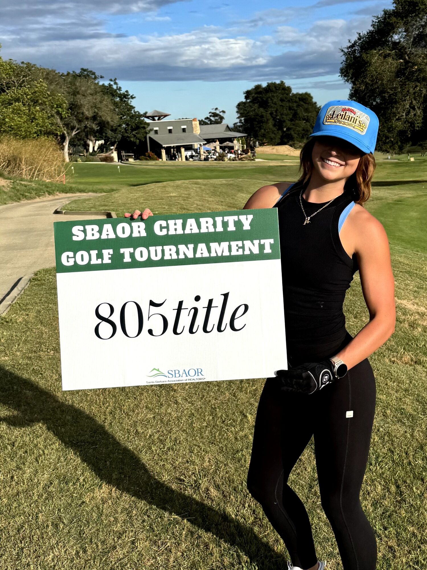 Woman holding a sign at a golf course, wearing a cap and sunglasses, with trees and a building in the background.