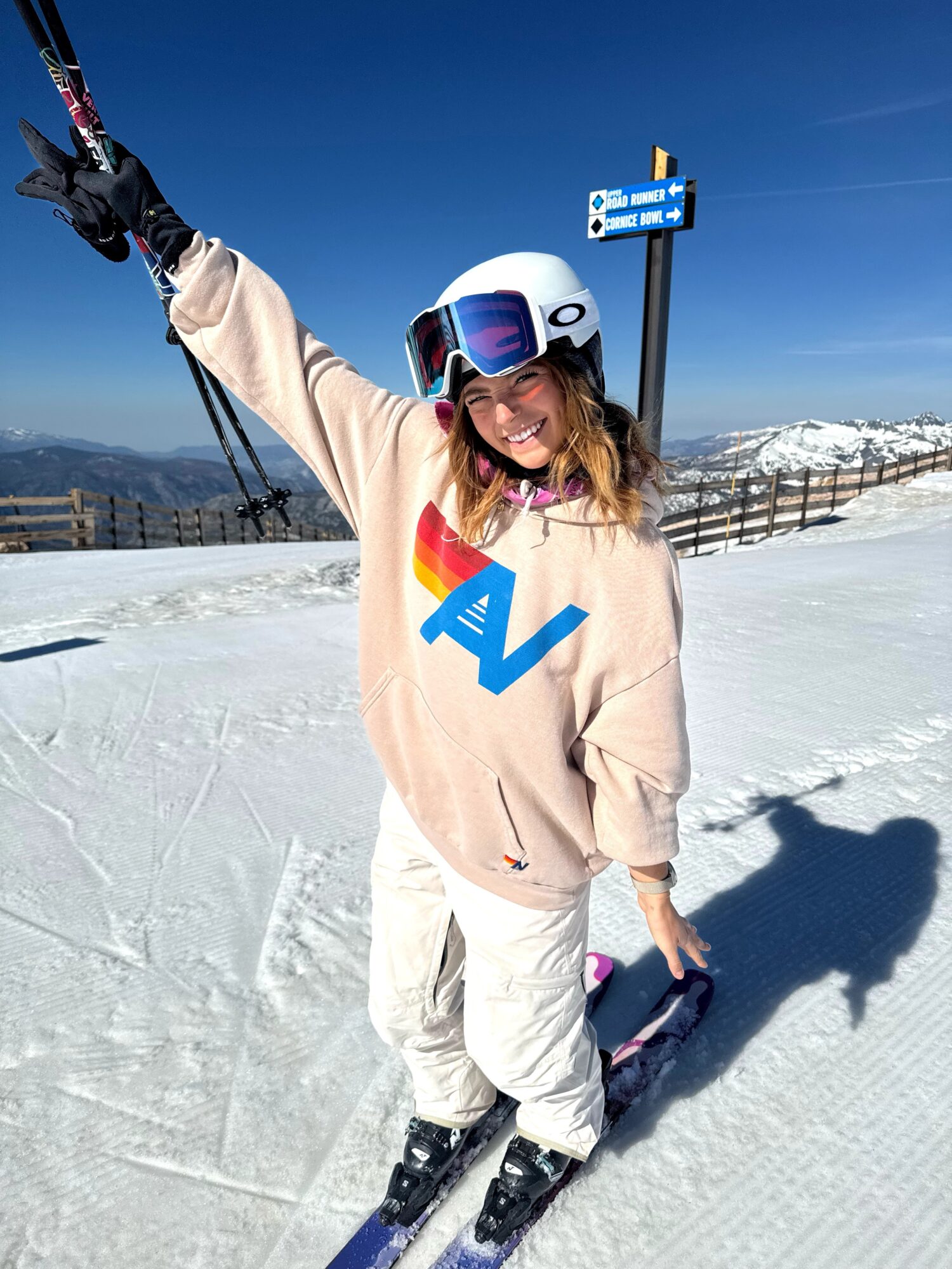 Person in ski gear smiling on snow, holding ski pole, wearing helmet and goggles, with mountains and blue sky in background.