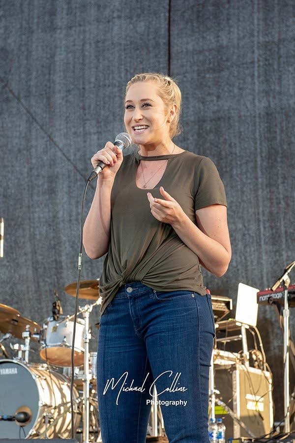 Woman holding microphone and smiling on stage with musical instruments in background.