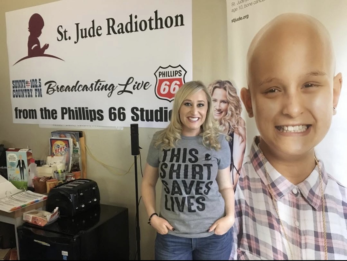 Young woman with blonde hair smiling, wearing a gray T-shirt with text, standing in front of a large poster of a smiling child, at a radiothon event.