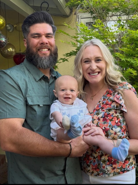 Three people, a man, a woman, and a baby, smiling outdoors with greenery in the background.
