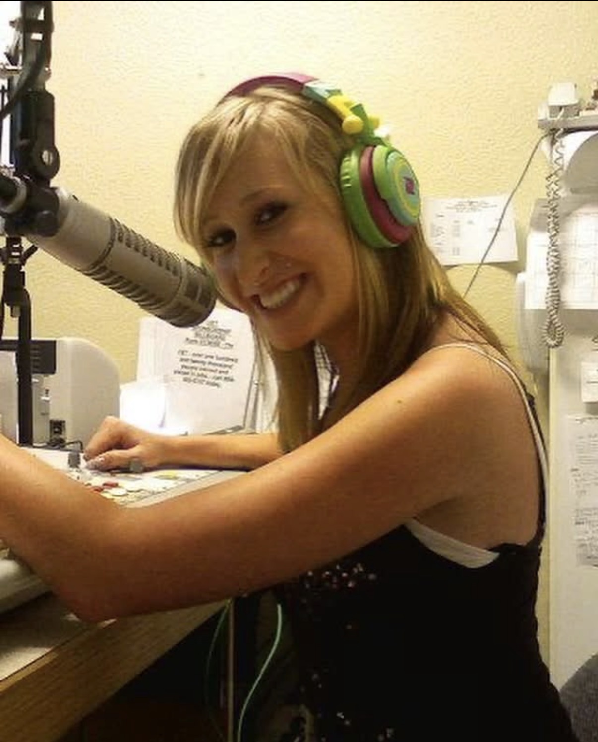 Woman with headphones speaking into a microphone in a radio studio, smiling, with equipment and papers around her.