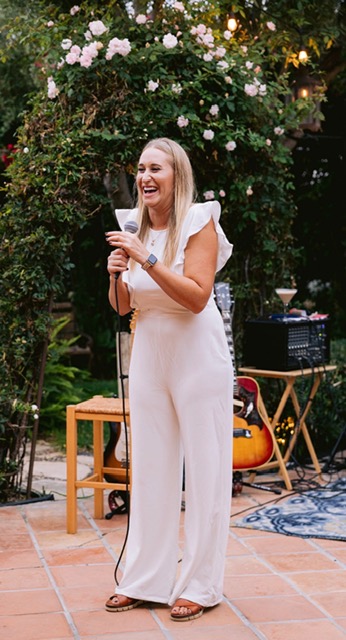 Woman in white jumpsuit holding a microphone, smiling outdoors with plants and a guitar in the background.