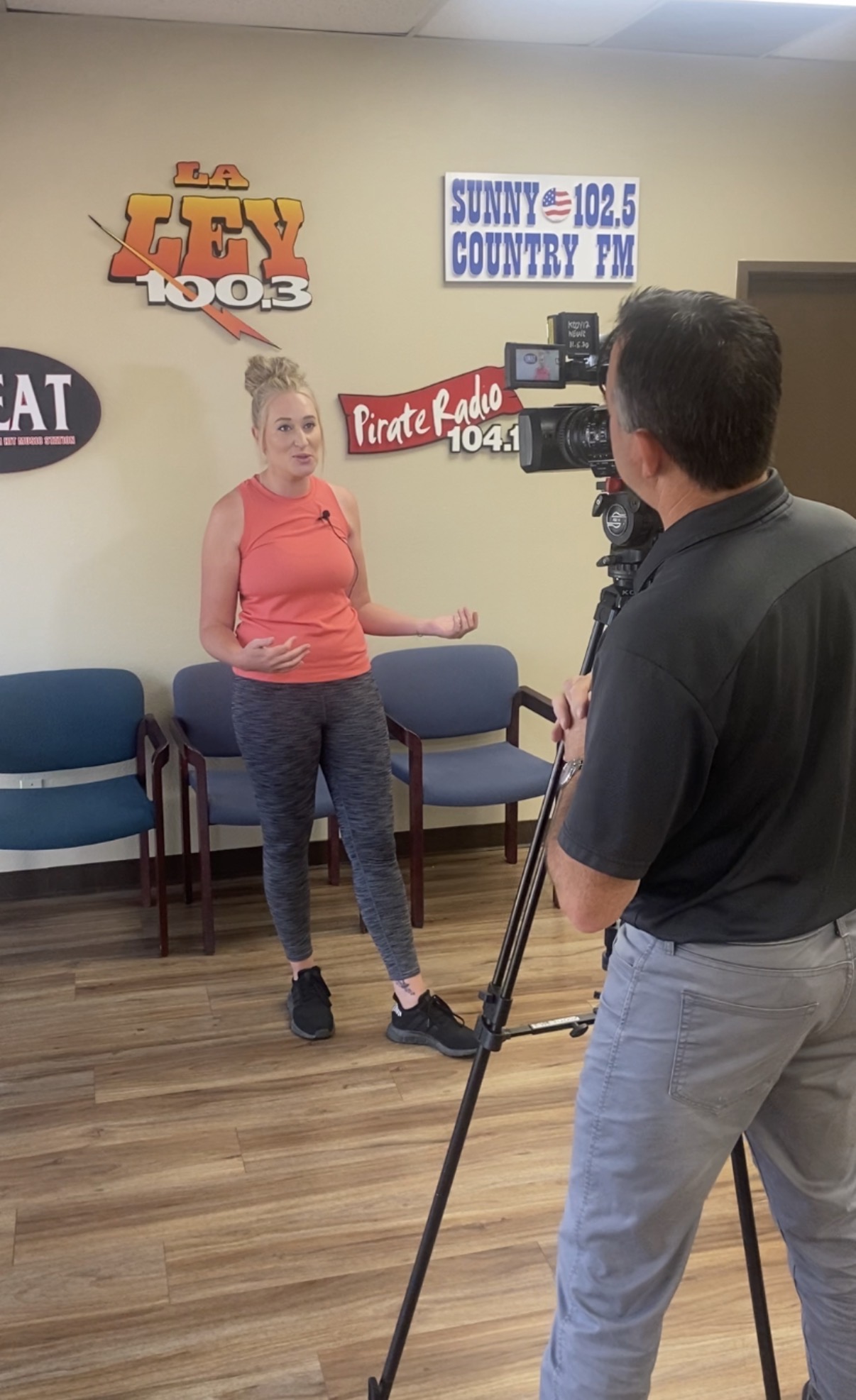 Young woman in red shirt and patterned pants being interviewed in a room with radio station signs on the wall.
