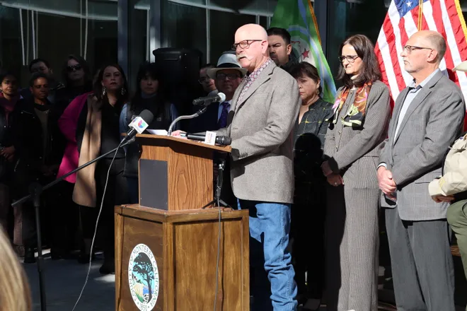 Man speaking at a podium during a public event with a group of people and flags in the background.
