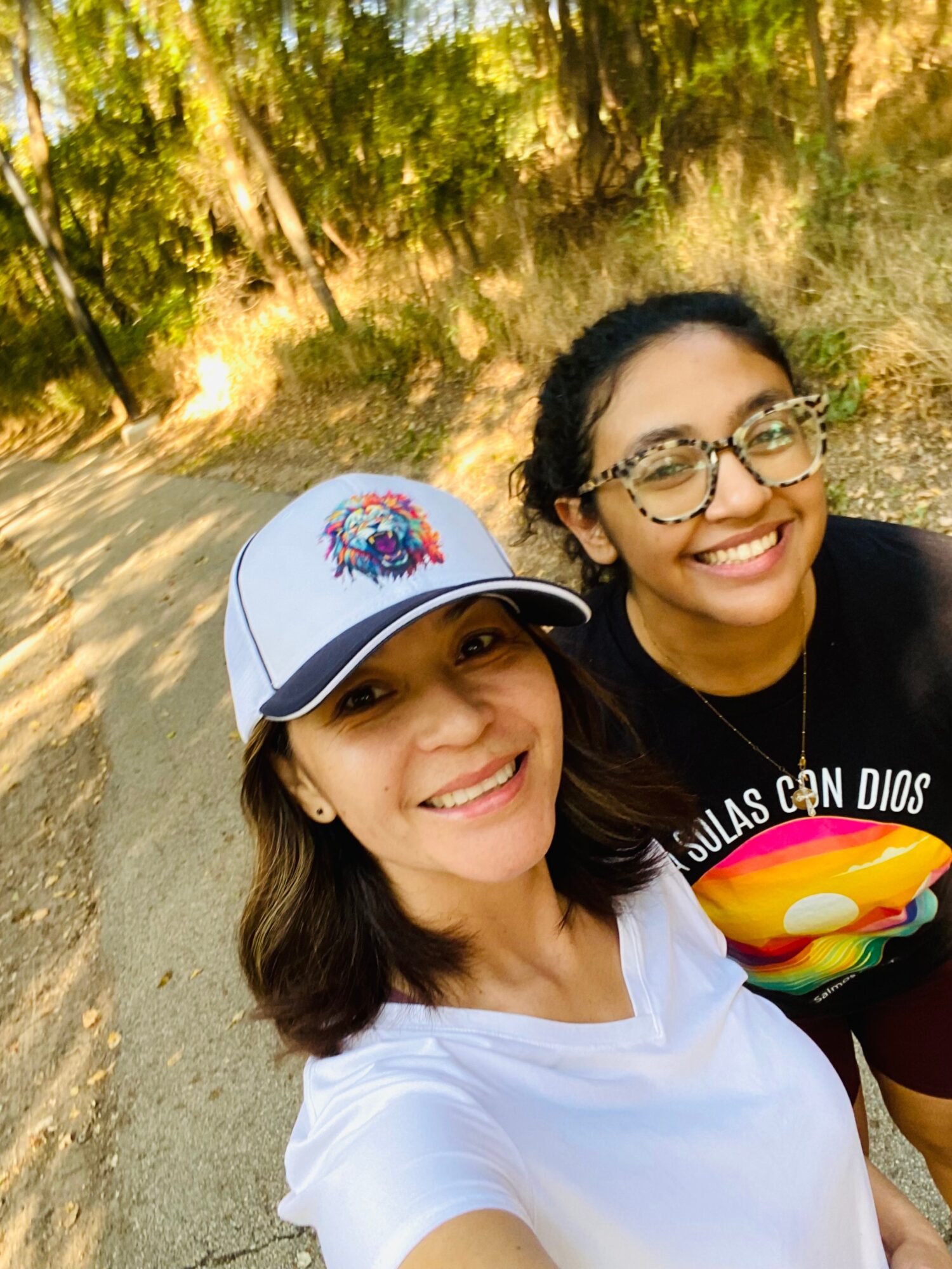 Two smiling women outdoors, one wearing a white cap and white shirt, the other with glasses and a colorful shirt, trees in background.