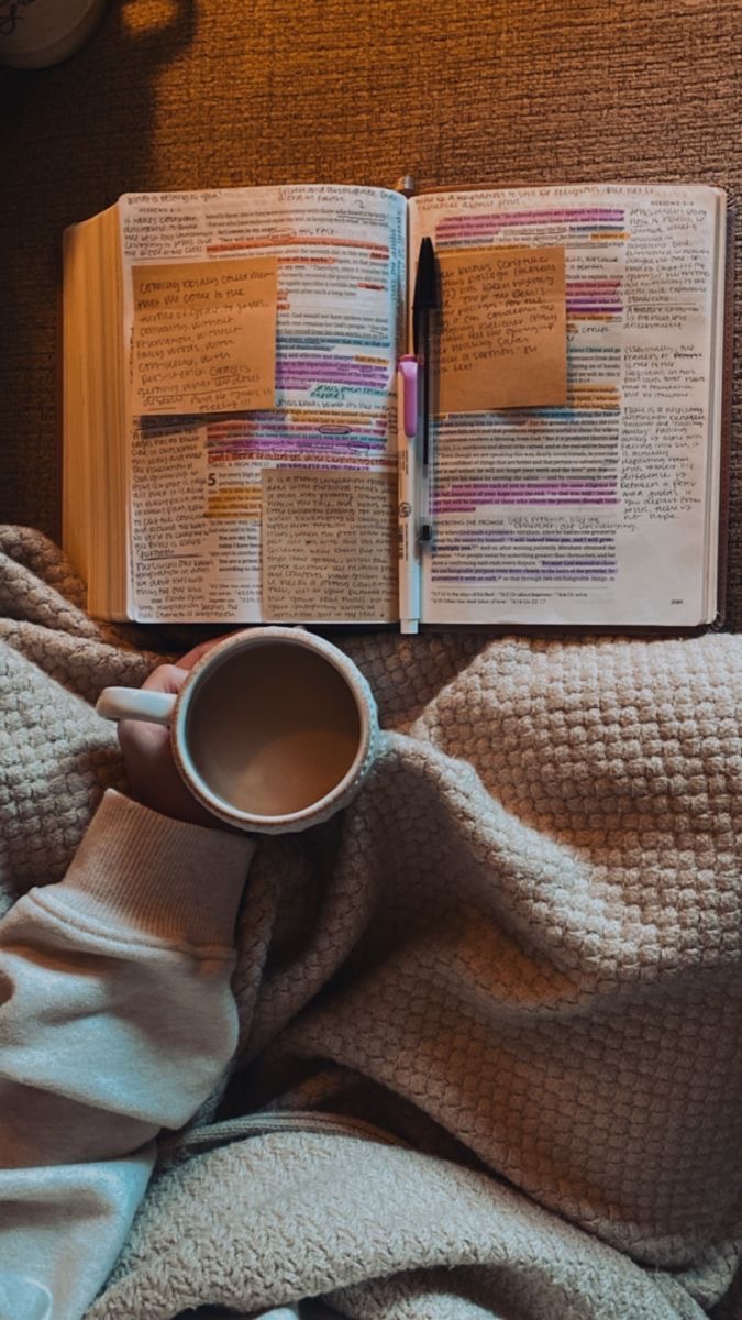 Open book with highlighted text and sticky notes, person holding a cup of coffee, cozy blanket, warm lighting.
