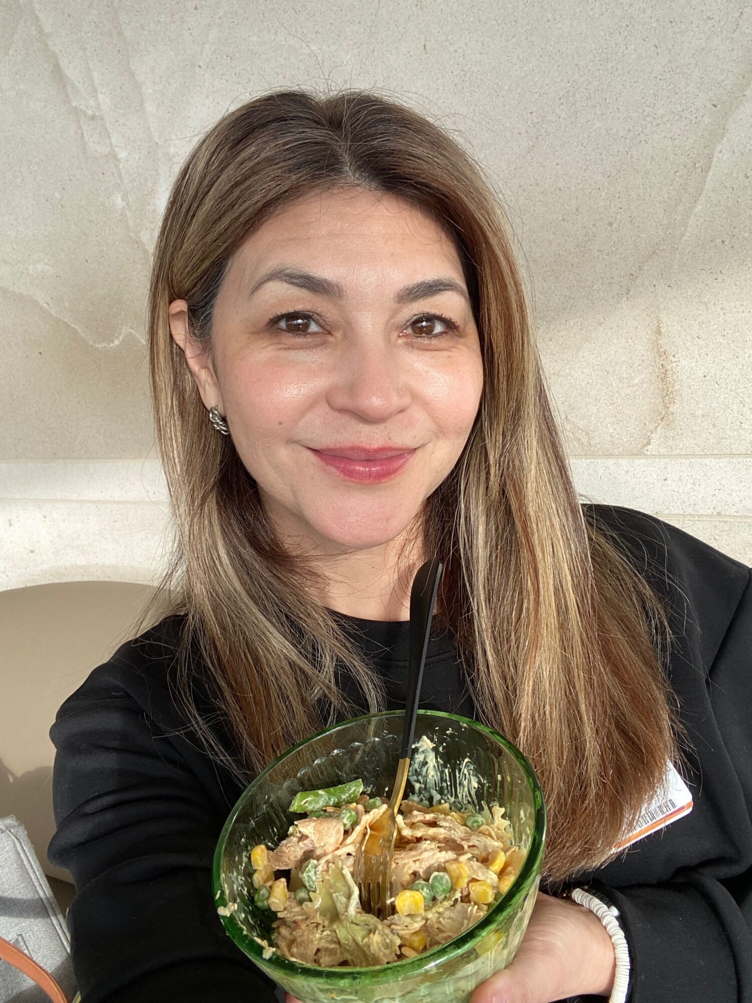Woman with long hair smiling holding a bowl of salad, wearing a black top, against a light background.
