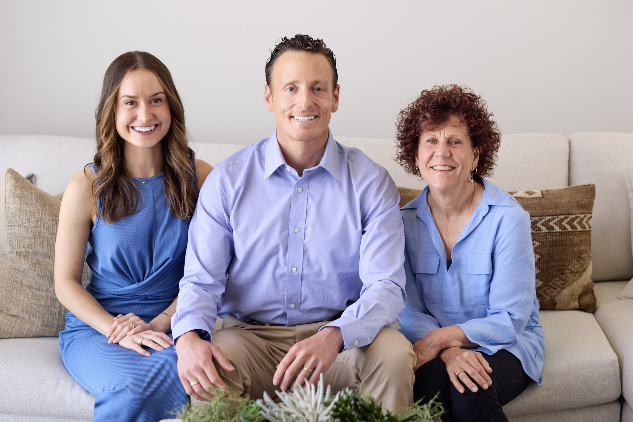 Three people sitting on a sofa, smiling, in a living room with pillows, wearing blue shirts and a dress.