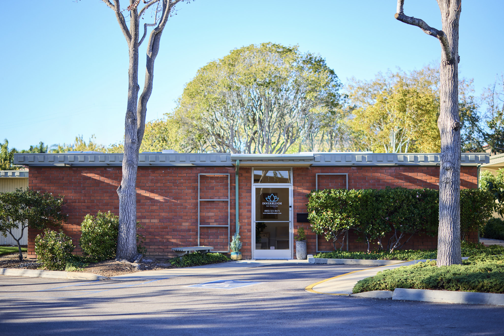 Single-story brick building with a glass door, surrounded by trees and bushes, under a clear blue sky.