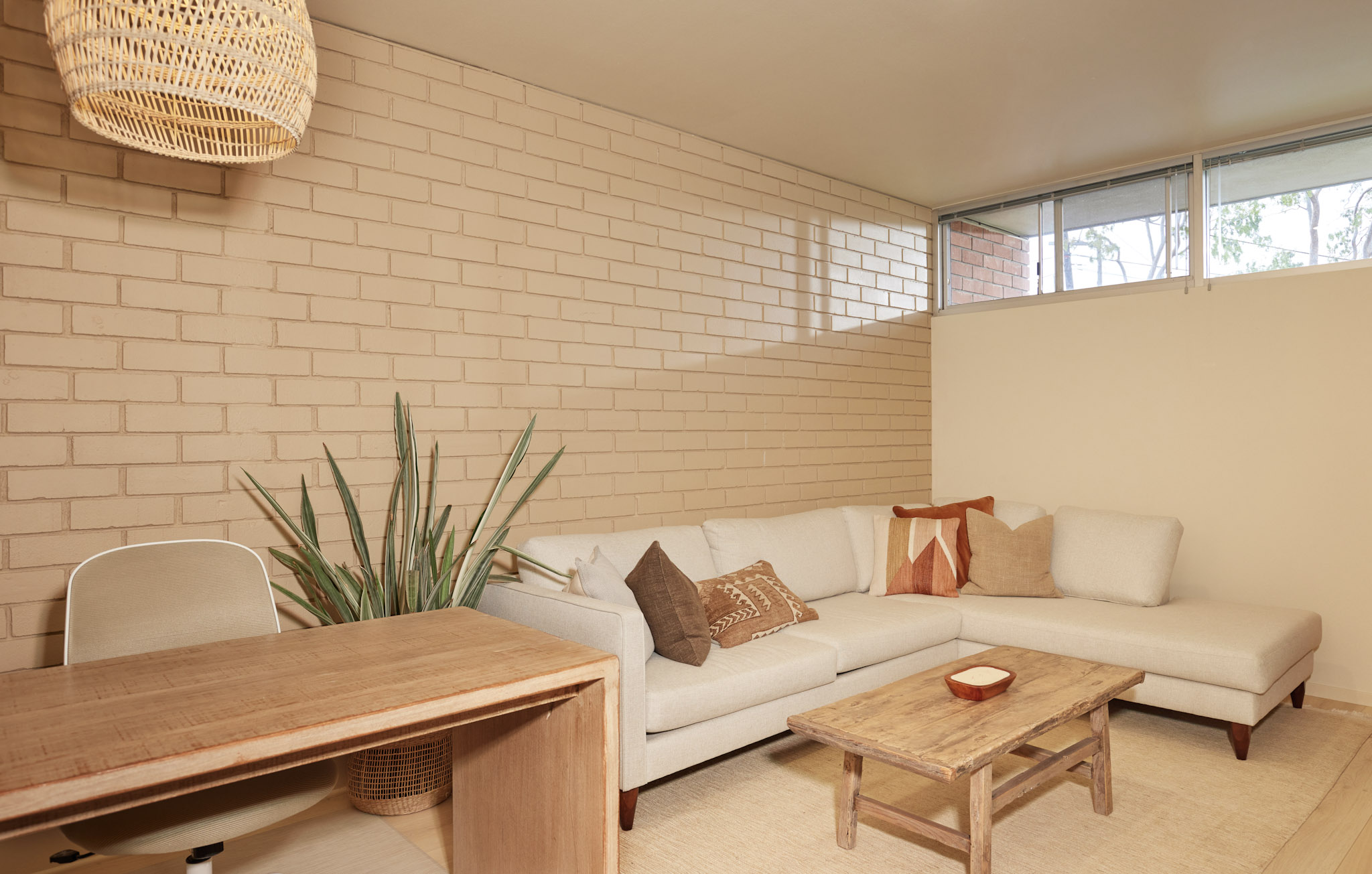 Living room with a white sectional sofa, wooden coffee table, and a plant, brick wall, and windows in the background.