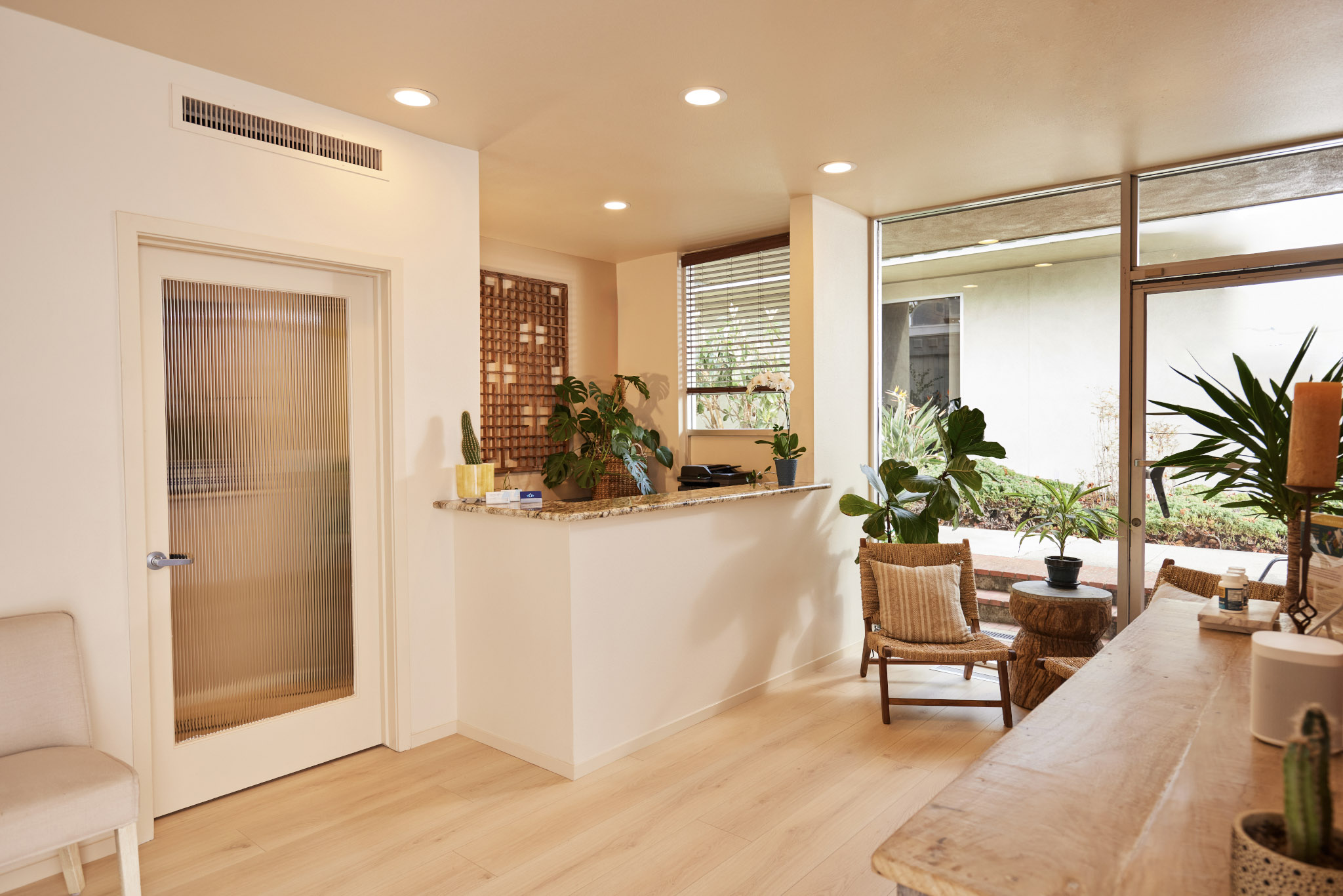 Bright interior with a reception desk, seating area, large window, and potted plants, with light-colored wooden flooring.