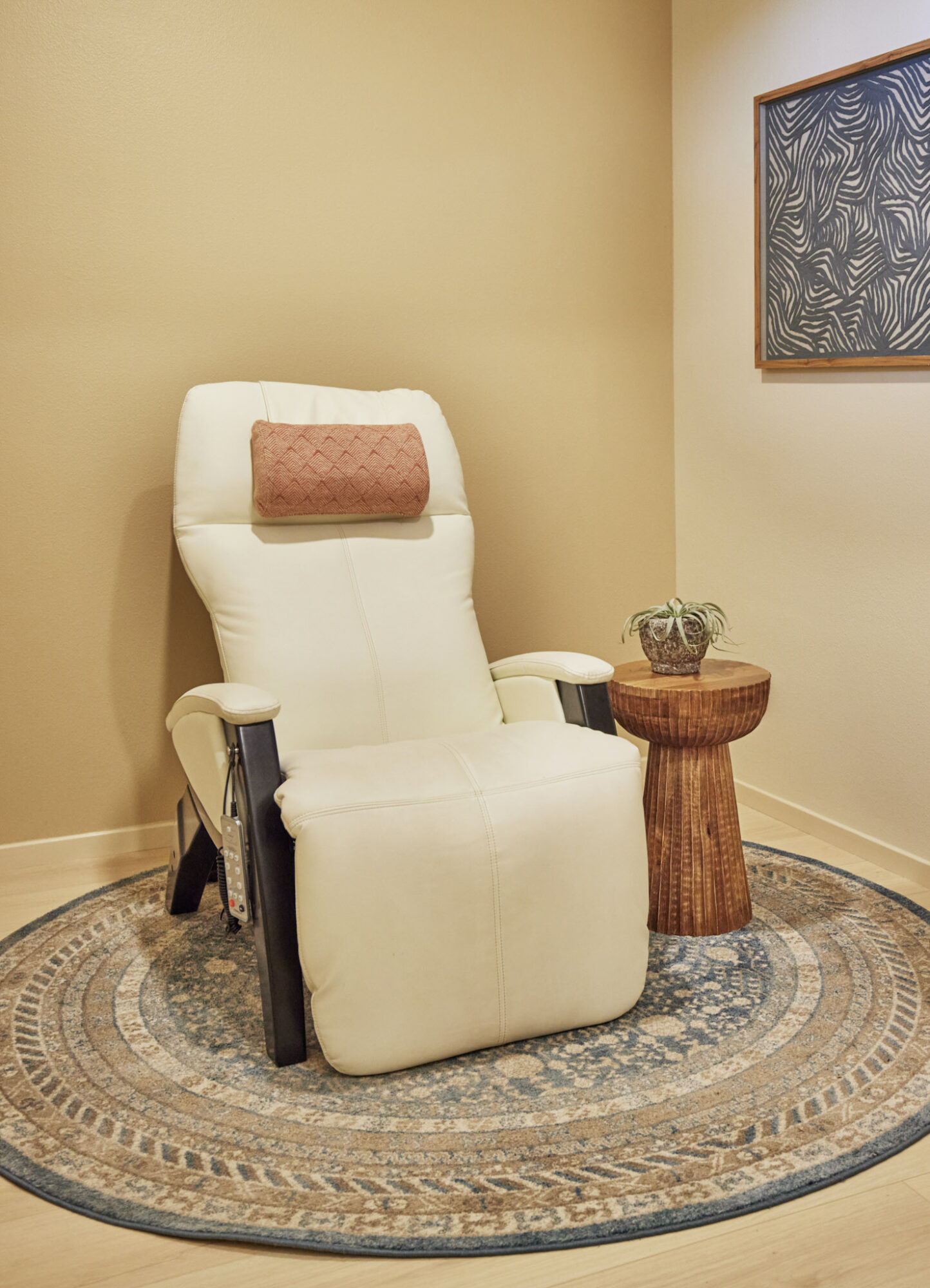 Recliner chair with a pink pillow, next to a small round wooden table with a plant, on a patterned rug.