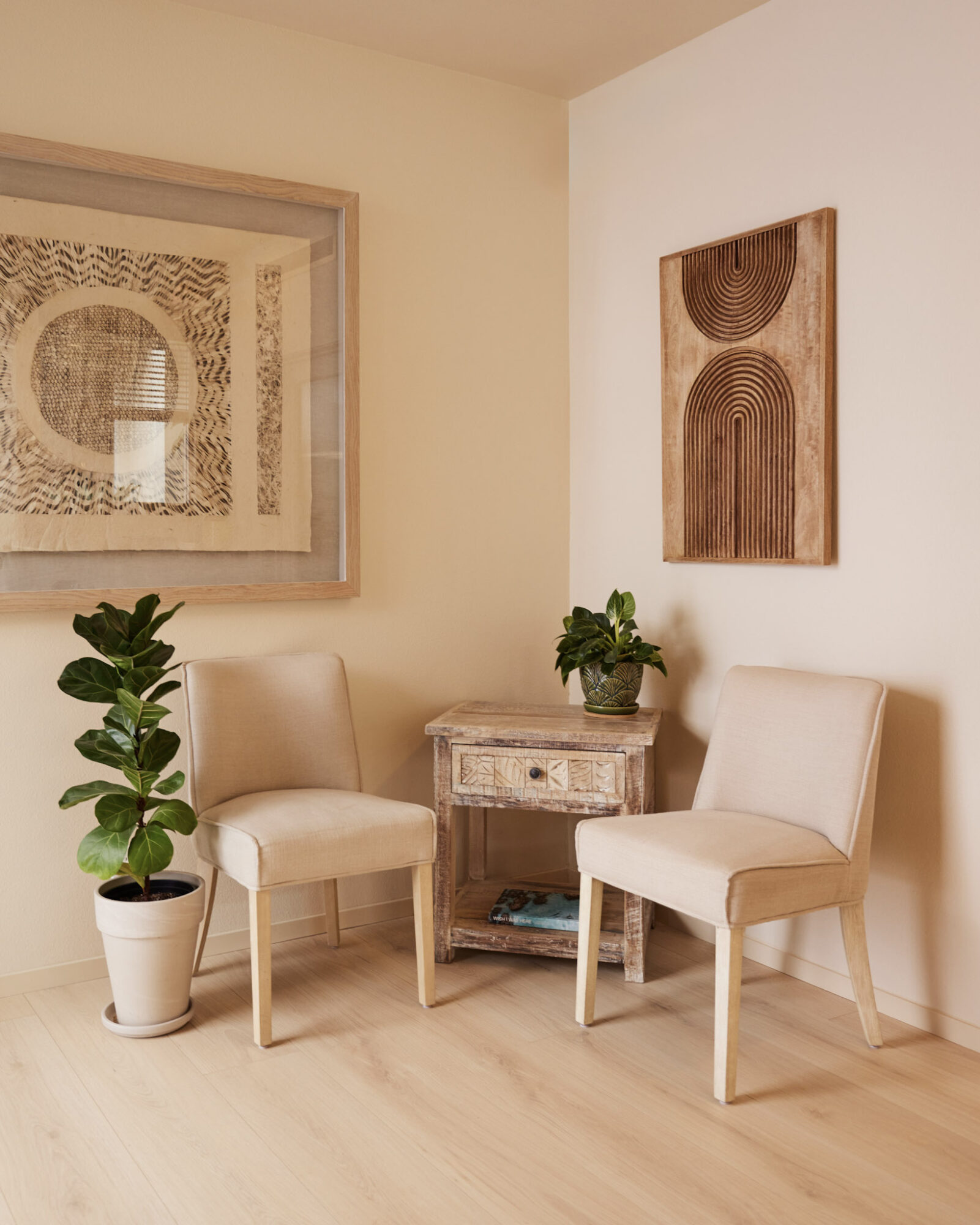 Two beige chairs with wooden legs, a small wooden side table with a potted plant, and artwork on the wall in a cozy room.