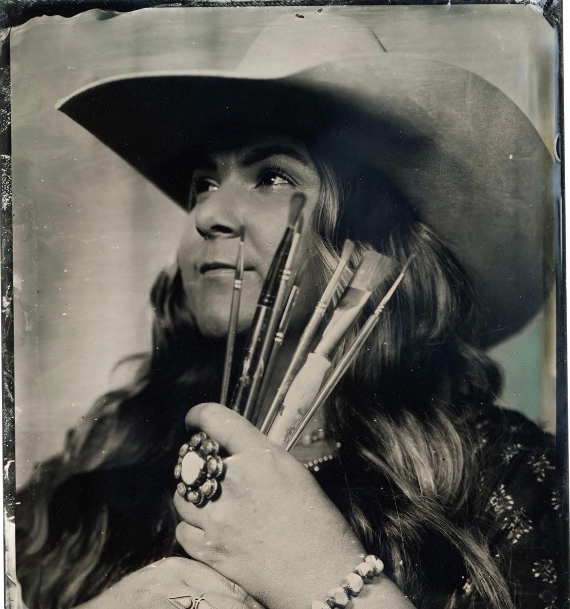 Woman wearing a large hat holding paintbrushes, with long hair and a ring on her finger.