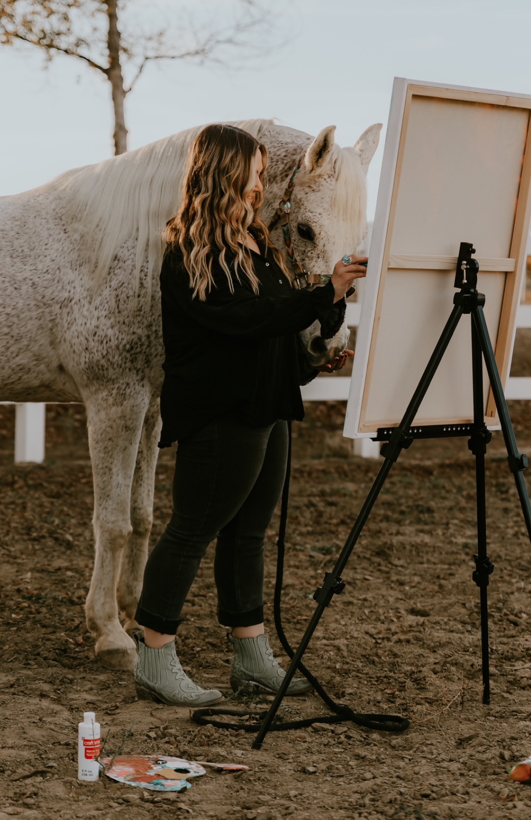 Person with a horse painting on a canvas outdoors, with art supplies on the ground.