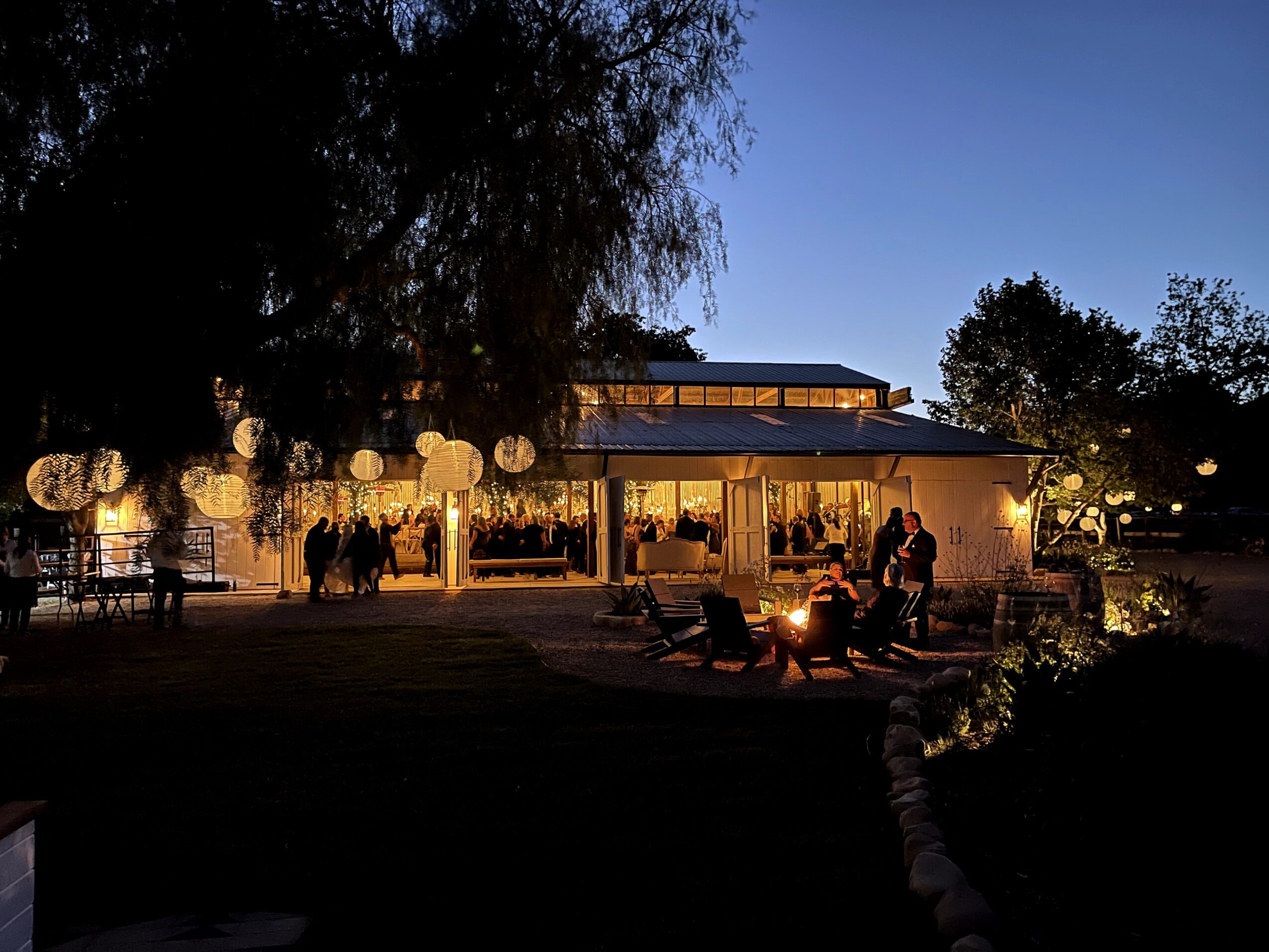 Nighttime scene of a modern building with large glass windows, outdoor seating, and people gathered around a fire pit.