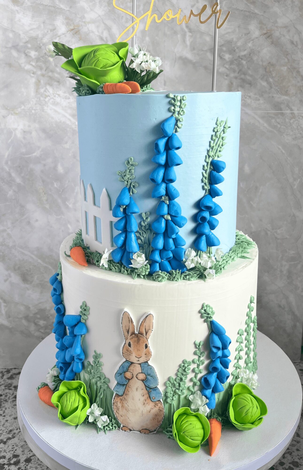 Two-tiered cake with blue floral decorations, a bunny illustration, and green and orange accents, against a gray background.