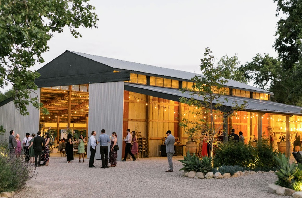 People gather outside a modern building with large glass windows and a sloped roof, surrounded by trees and landscaping.