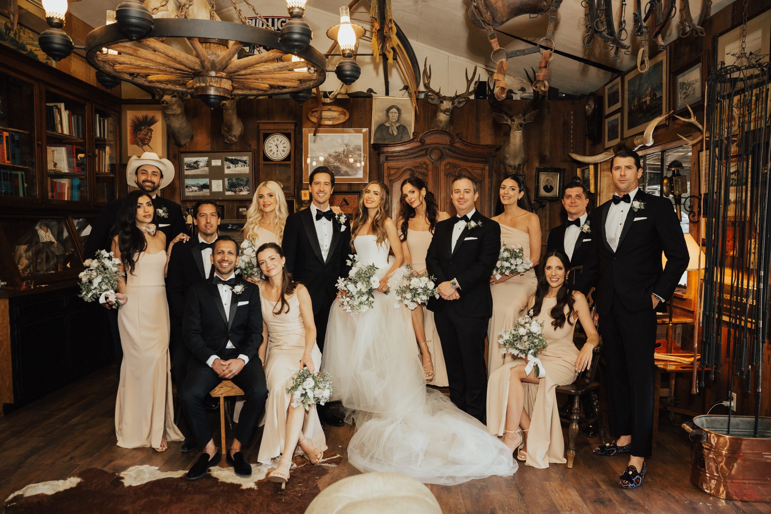 Group of people in formal attire, some holding bouquets, in a vintage-style room with wooden furniture and decorative items.